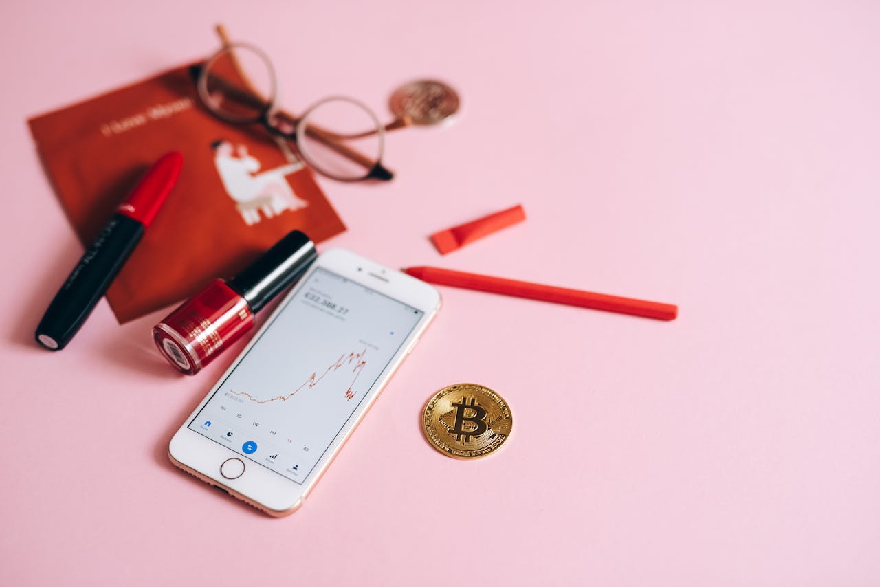A flat lay of a smartphone with Bitcoin, makeup items, and glasses on a pink surface, illustrating finance and fashion.